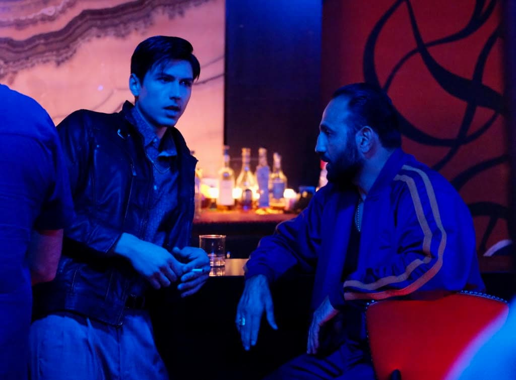 Roman Mitichyan in a blue tracksuit talking to another actor across a table in a neon-lit bar scene