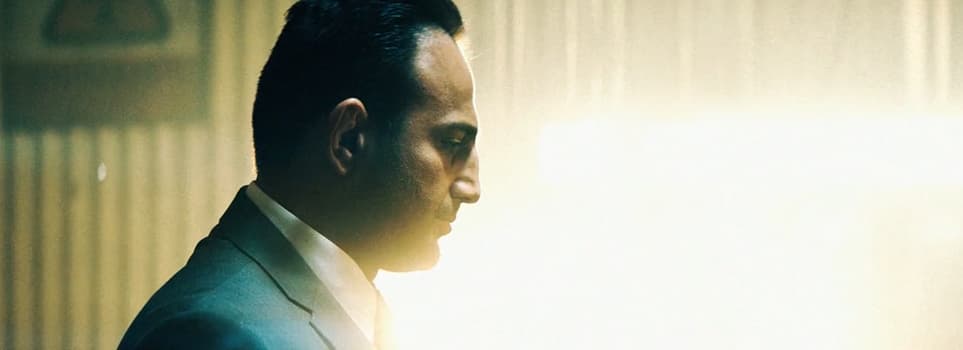 Roman Mitichyan in a dark suit, seen in profile in a warehouse in Extraction